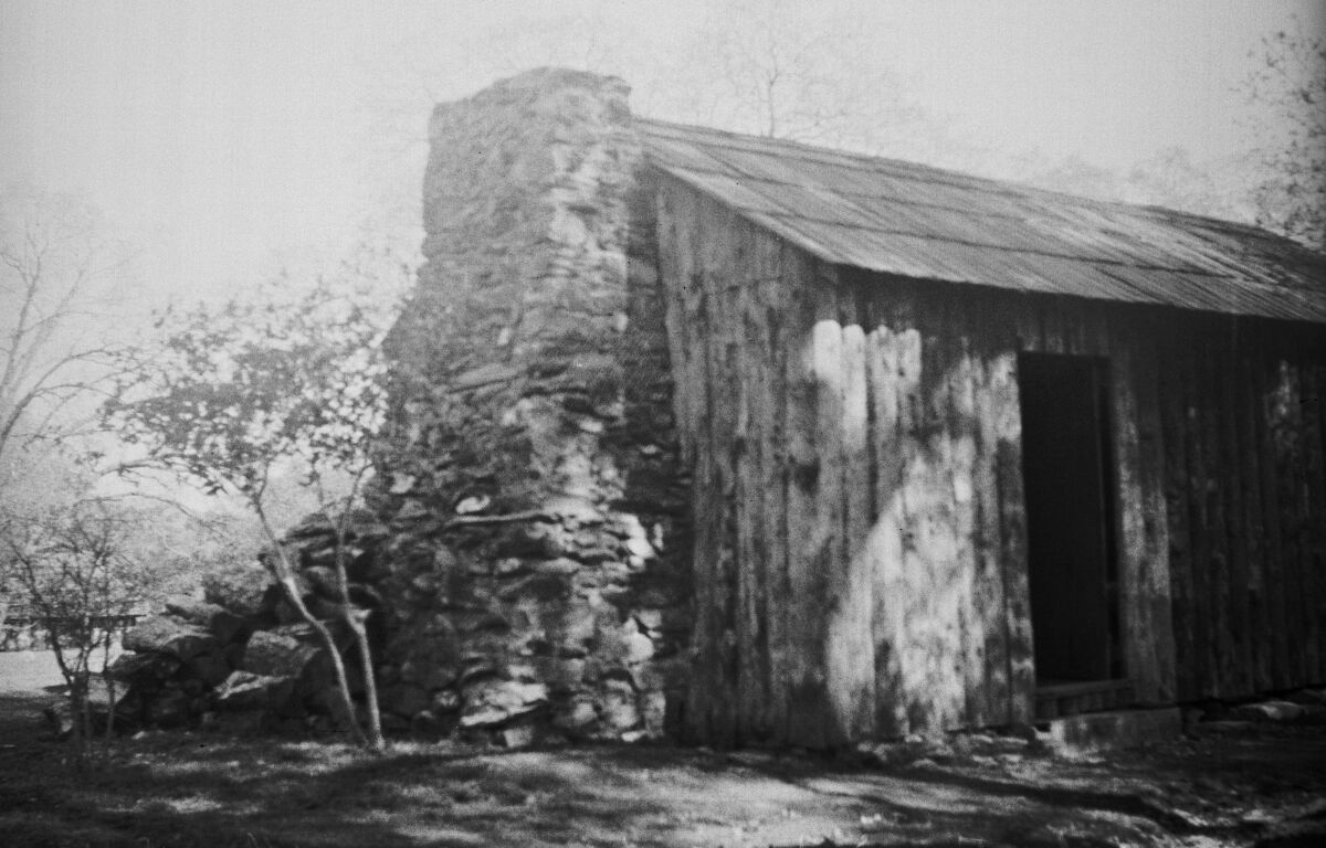 A black-and-white photo of a rustic log cabin with a rock chimney and no windows.