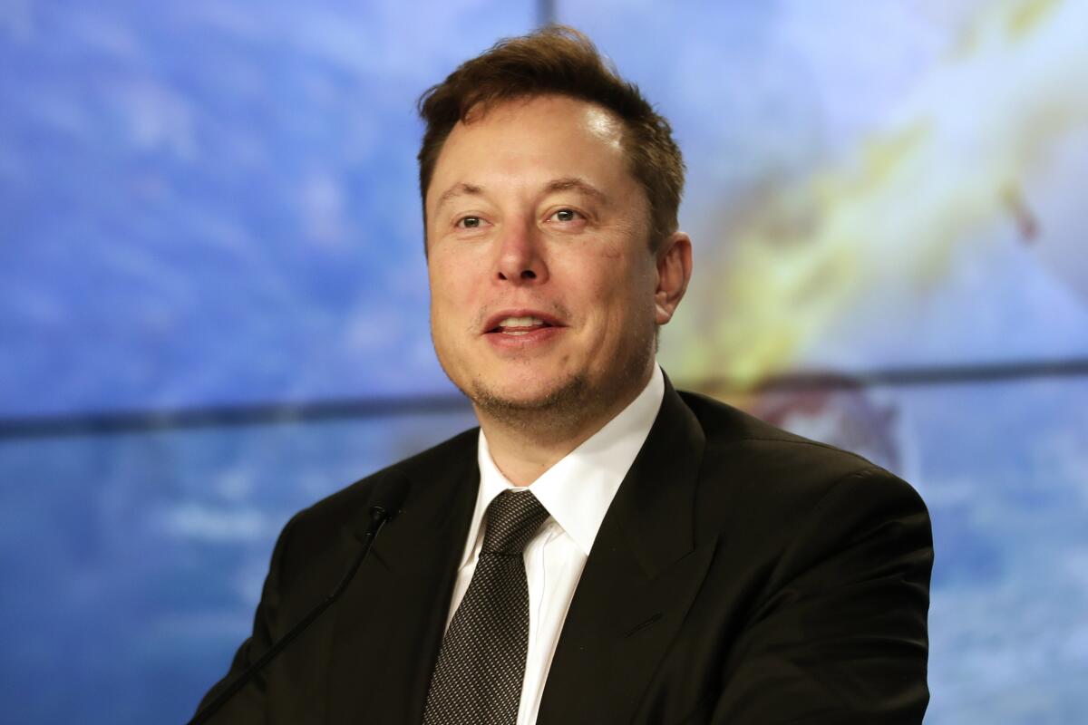 Elon Musk in a black suit and tie sitting in front of a blue screen.