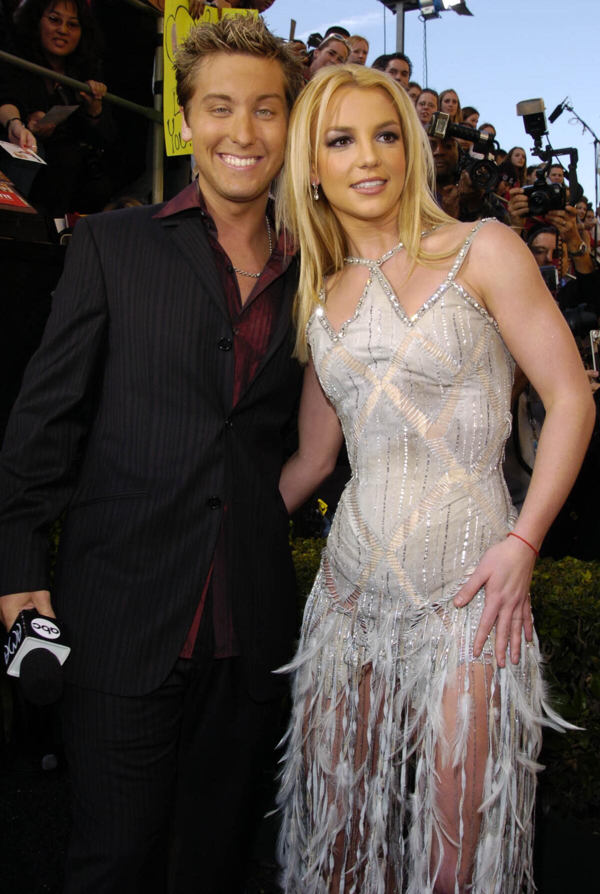 Lance Bass is dressed in a black suit and is posing together with Britney Spears who is wearing a silver dress