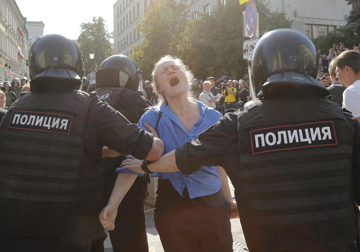 Two Russian officers wearing bulletproof vests and helmets detain a woman whose head is back and mouth is open as if in a yell.