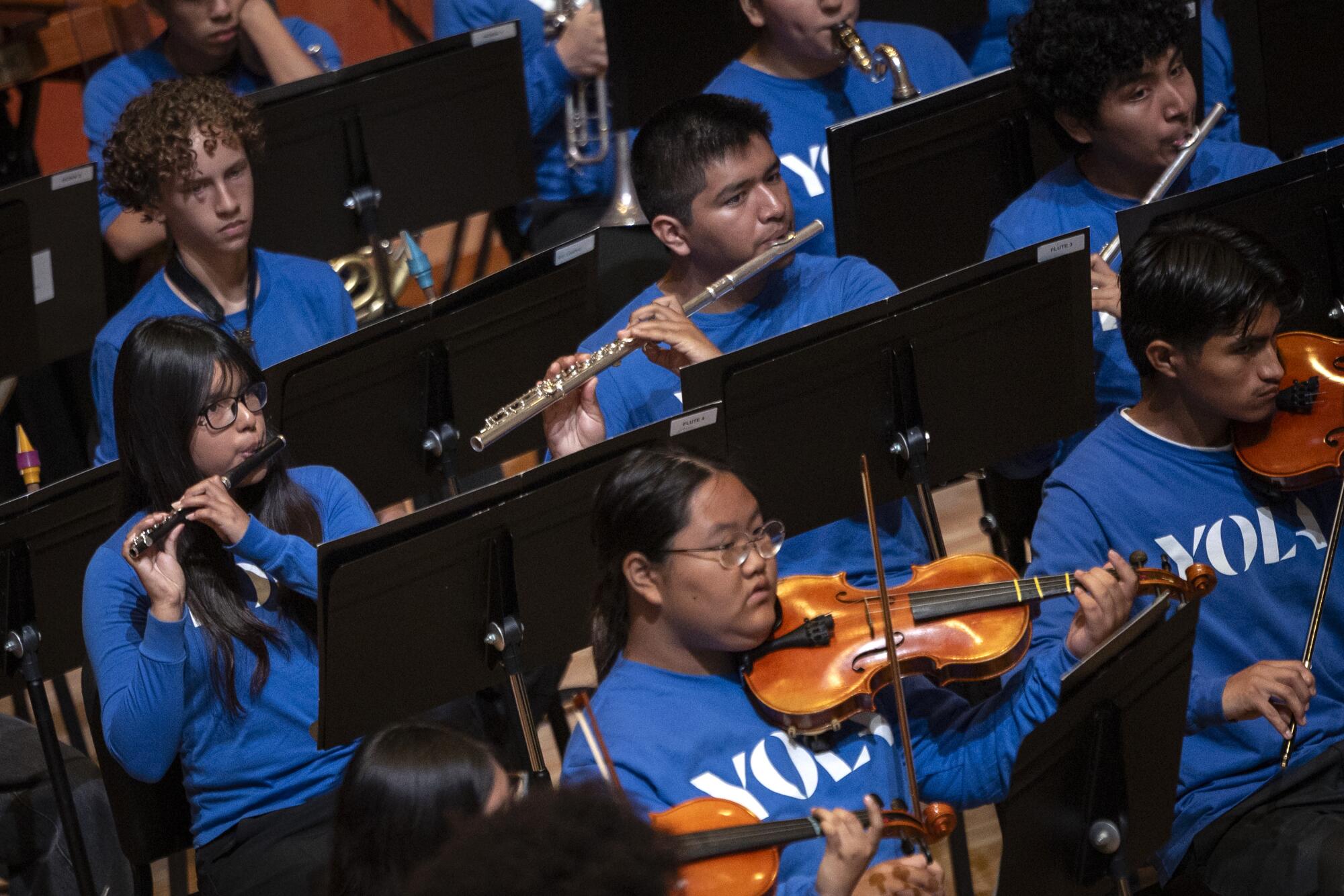 Crianças, vestindo camisas azuis da YOLA, tocam instrumentos em uma orquestra.