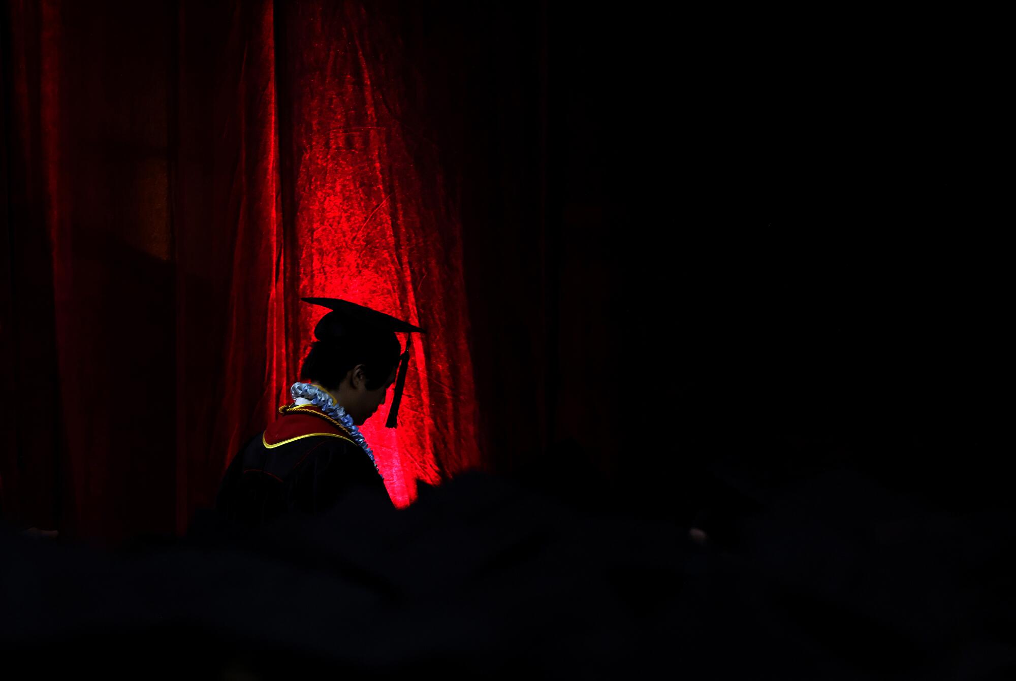 A USC student in graduation attire is partially silhouetted near a red curtain.