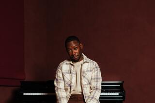 Lamorne Morris sits on a piano bench in a cream flannel and brown pants, with a red backdrop during a 2024 portrait session