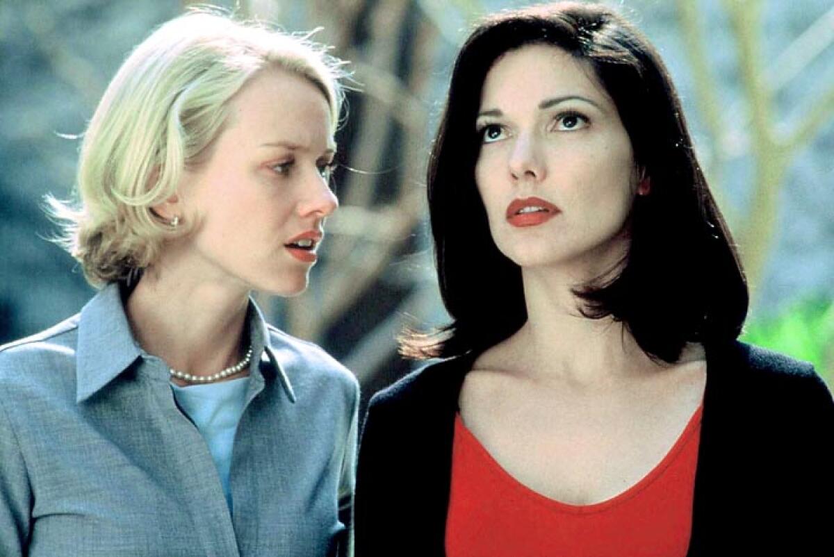 A blonde woman on the left wears a blue blouse and a pearl necklace and a brunette woman on the right wears a red blouse.