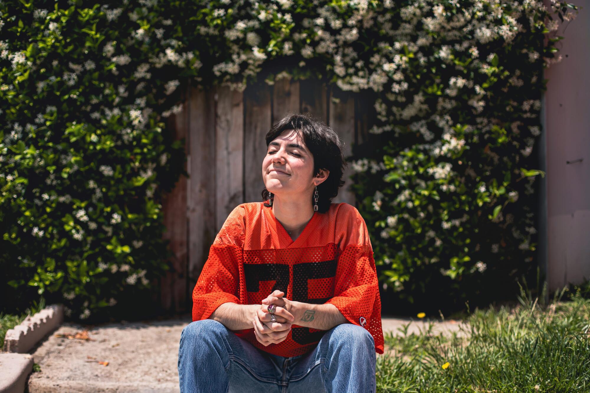 A person in red shirt and jeans sits outdoors in the sunshine, eyes closed, looking up