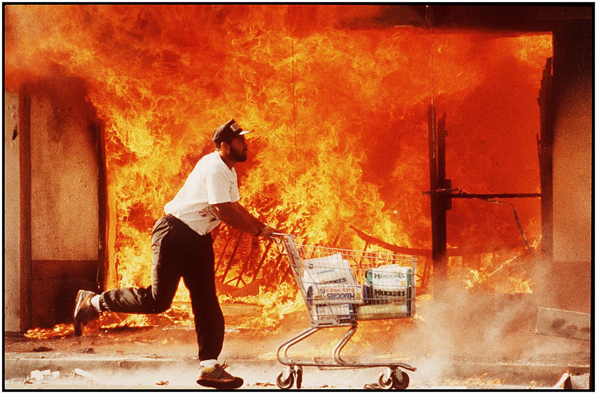 A man pushes a shopping cart. Flames burst from a building in the background.