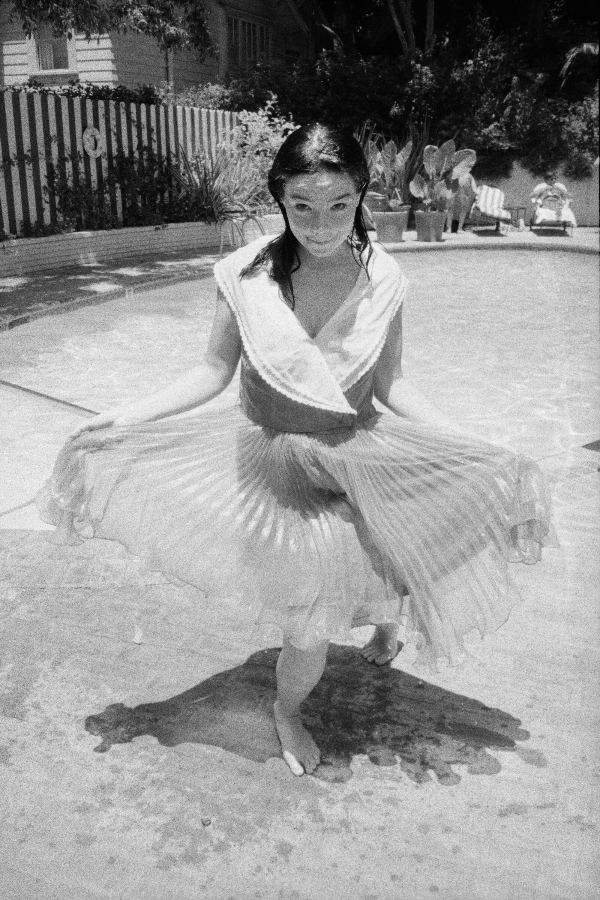 A black-and-white photo of Björk holding up the edges of her dress as she stands in front of a swimming pool.