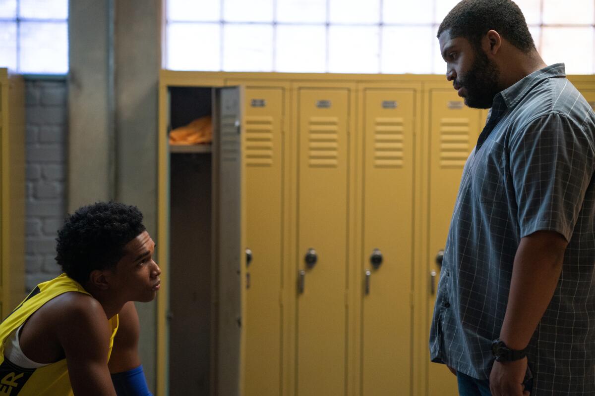 A teen basketball player looks up at his coach from a locker room bench.