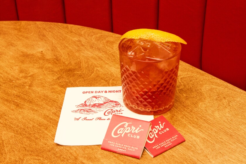 A photo of a negroni in a crystal tumbler, garnished by orange slice, sitting next to branded matchbooks and napkin.