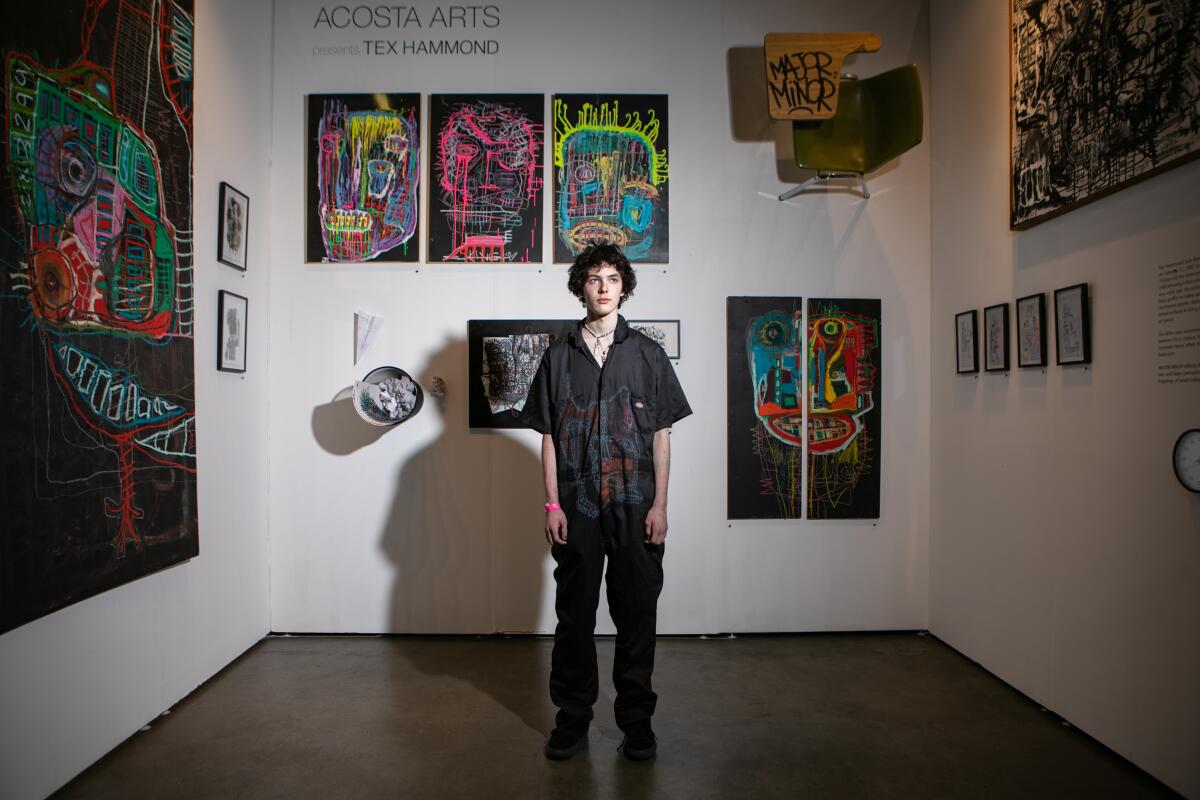 A teenager stands in a small room where his paintings are displayed on the walls.