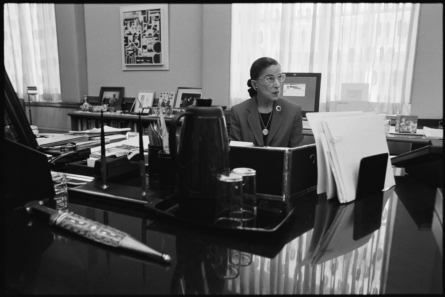 Black and white photo of Ruth Bader Ginsburg seated in front of her desk in her Supreme Court chambers