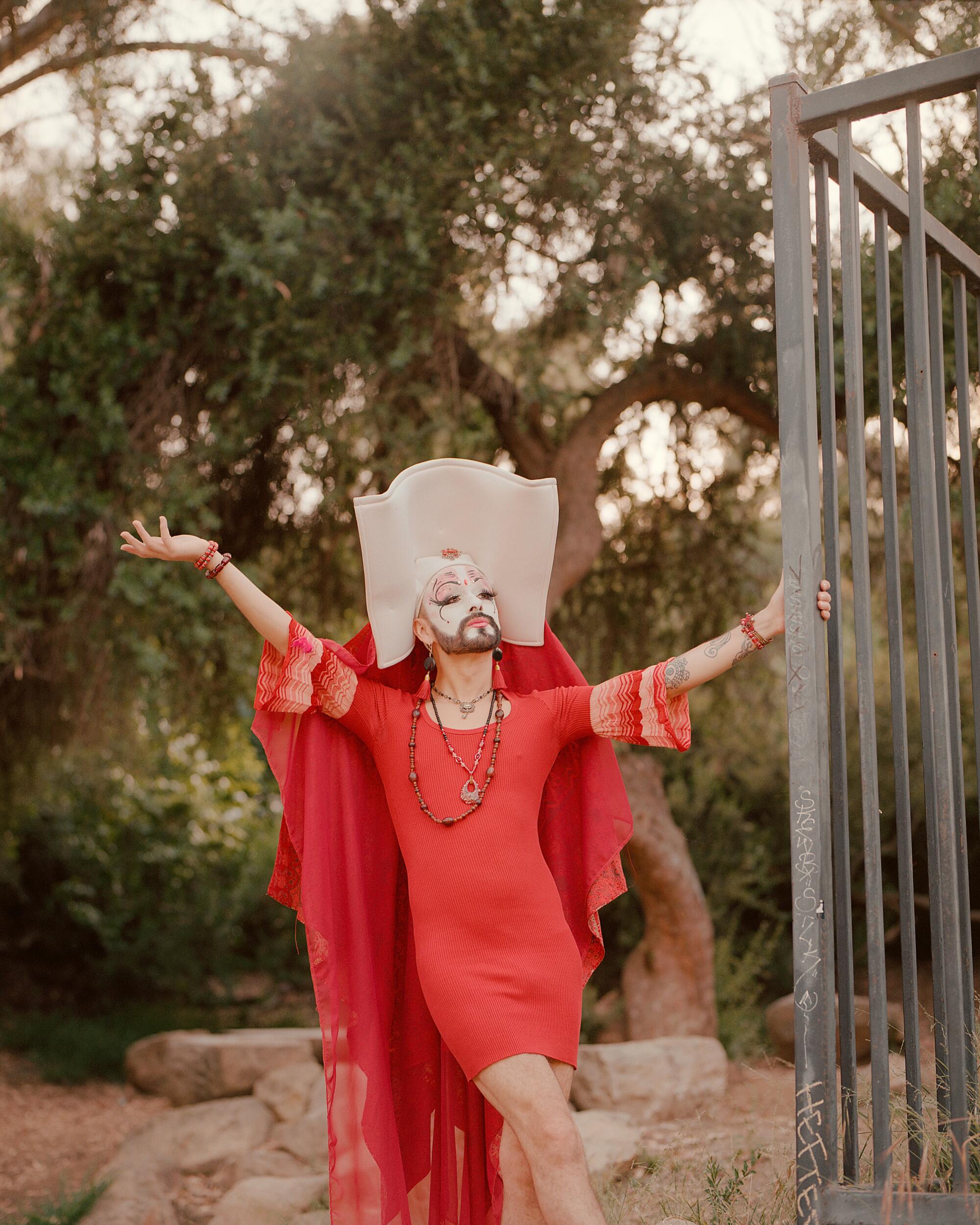A drag queen in a red dress, beard, face paint and ornate wimpole headdress strikes a pose outdoors.