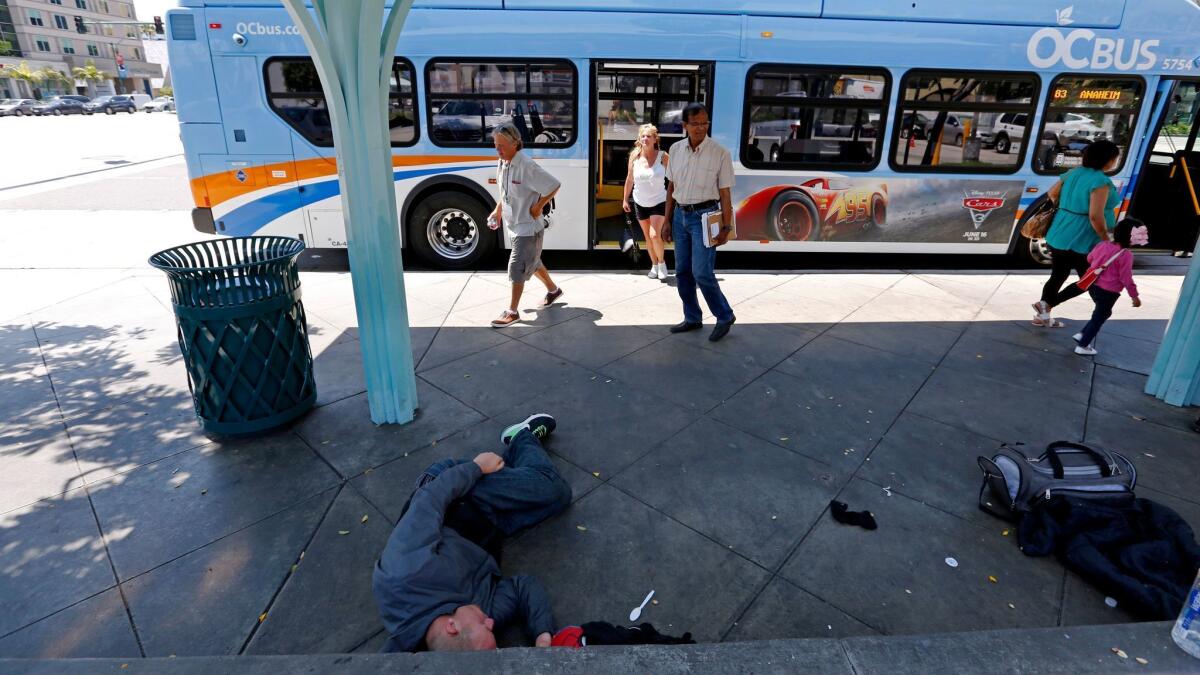 Un hombre desamparado duerme debajo de un refugio de autobús mientras la gente sale de un vehículo de la Autoridad de Transporte del Condado de Orange, en Harbor Boulevard y Katella Avenue, en Anaheim (Gary Coronado / Los Angeles Times).