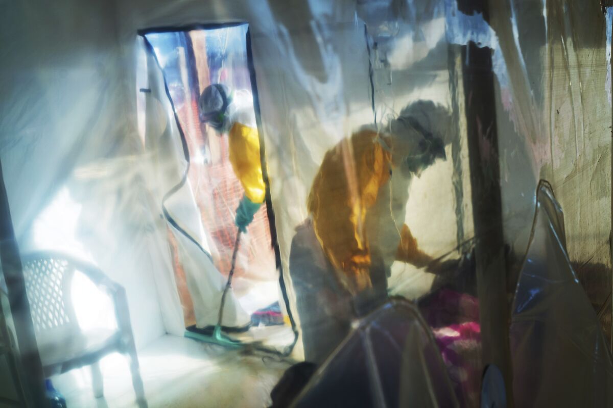 A person in a protective suit stands over a person on a bed inside a transparent tent.