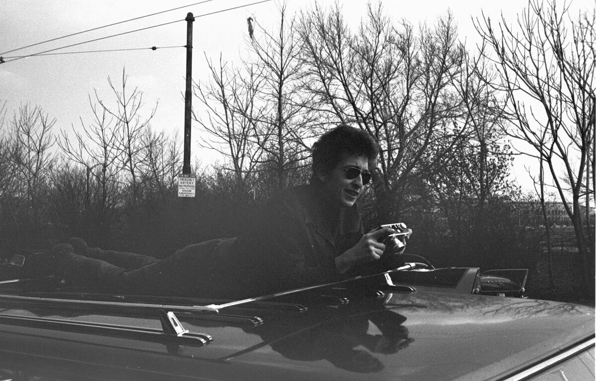A black and white photo of Bob Dylan lying on top of a convertible in 1964