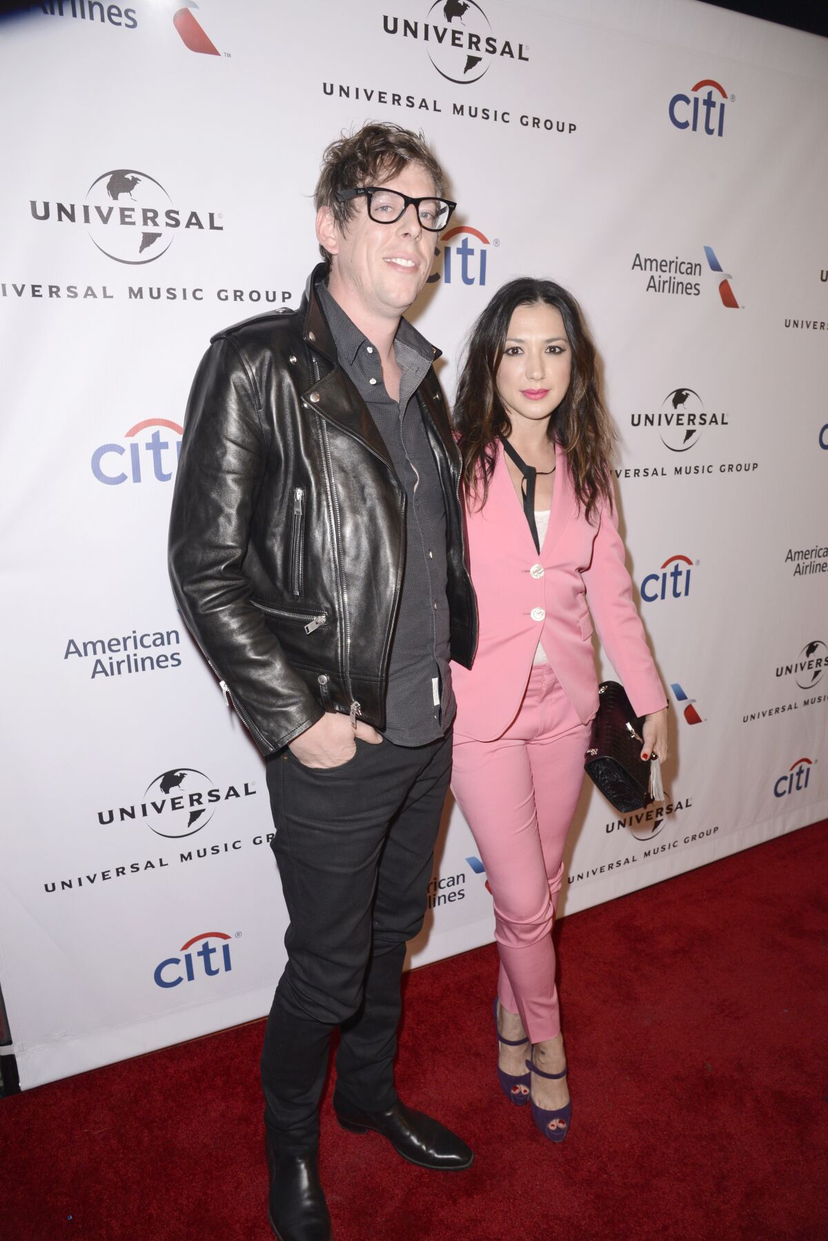 A man with glasses and a leather jacket and a woman in a pink suit pose on a red carpet