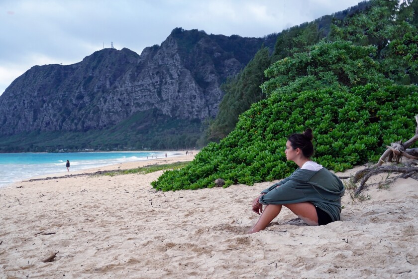 A teen girl sits on a beach in Hawaii, with high cliffs in the background