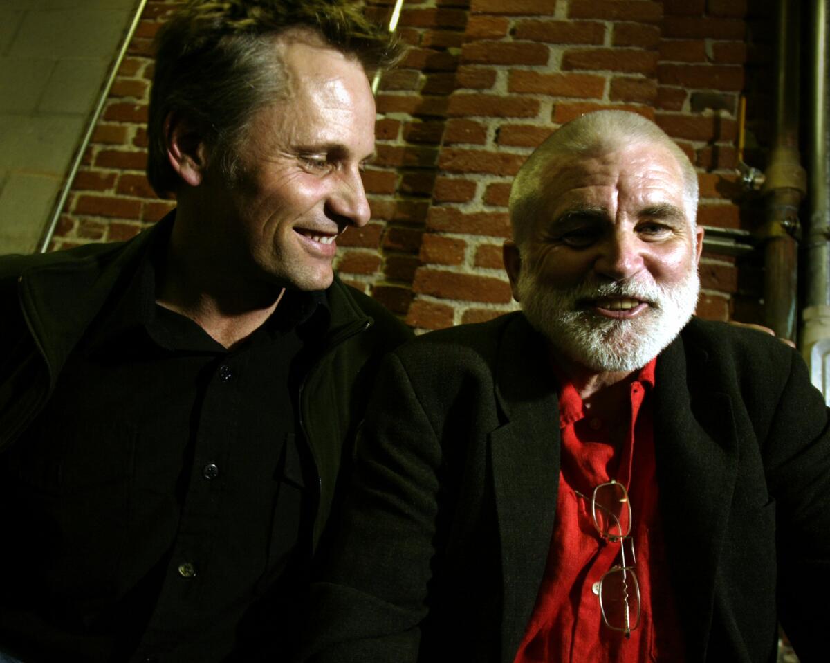 Mike Davis, in a red shirt and blazer,  smiles as Viggo Mortensen looks on.