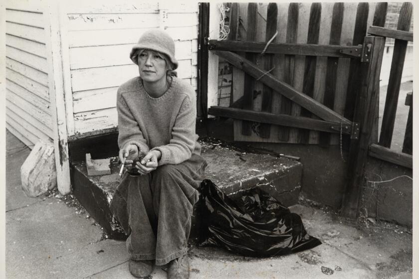 Eve Babitz in a hat sits and smokes a cigarette.
