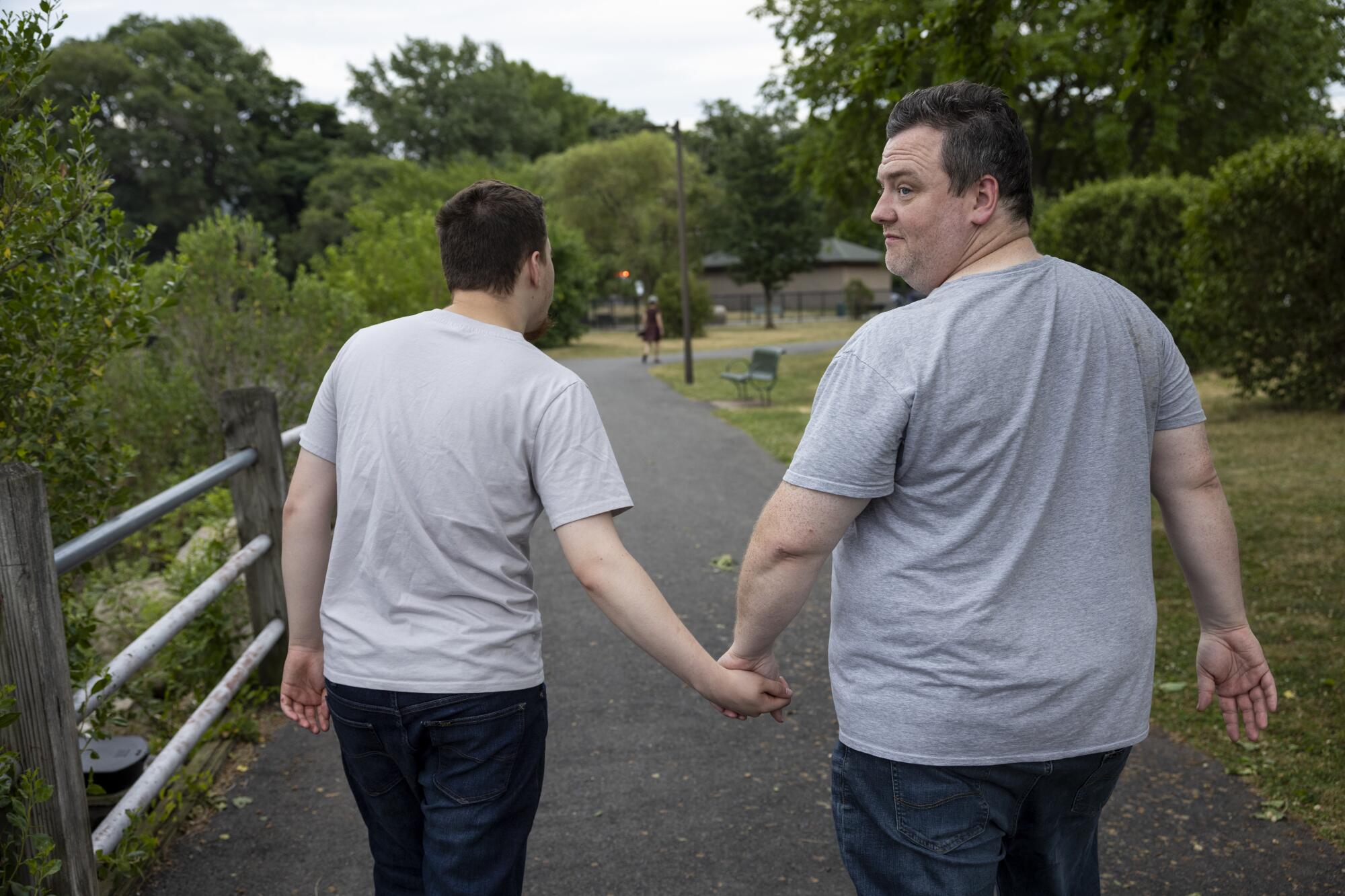 A man holds his son's hand in a parklike setting. They're walking away from the camera, the man looking back. 