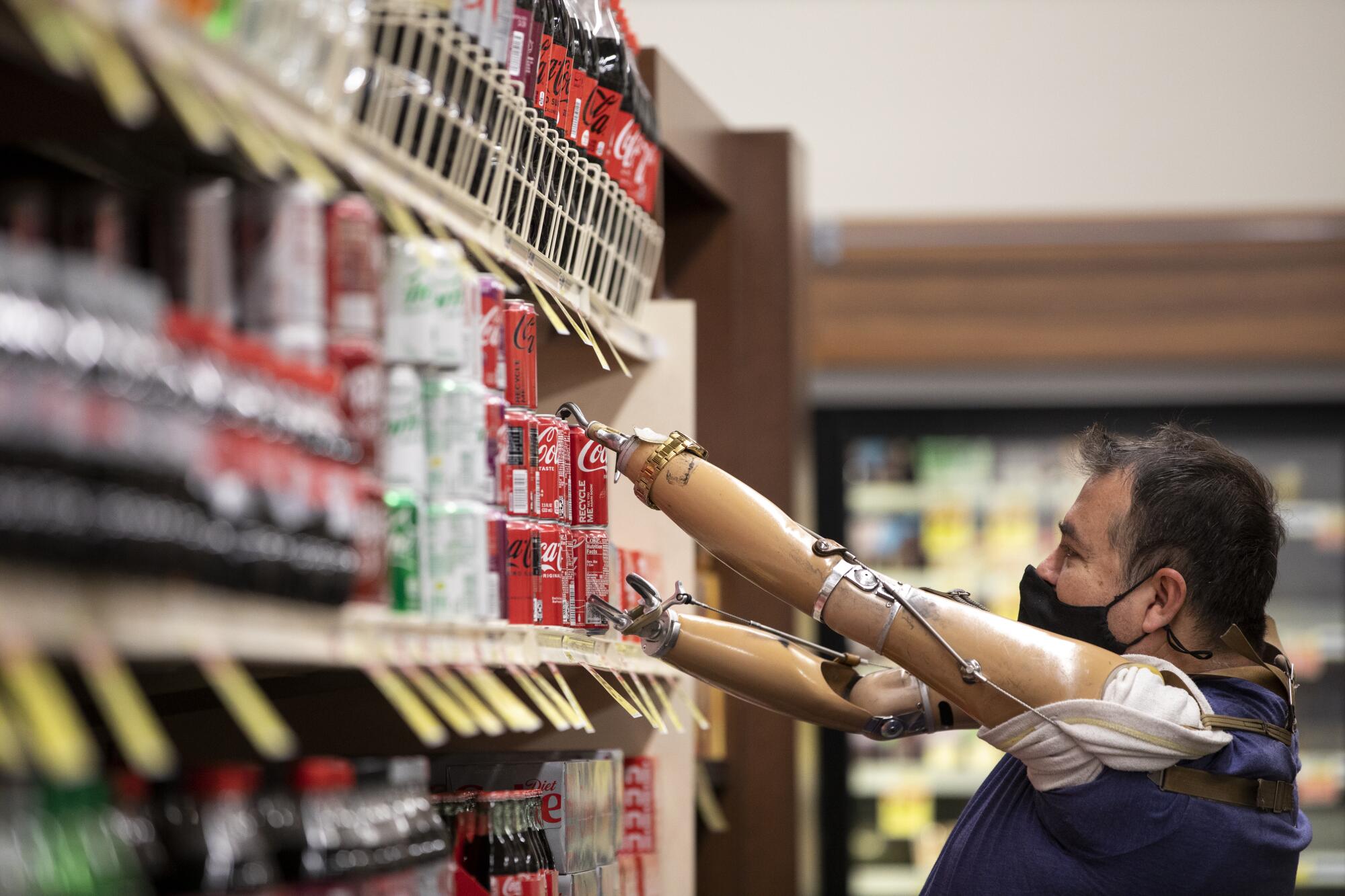 a man with two prosthetic limbs wearing a face mask reaches for a six pack of Coca Cola