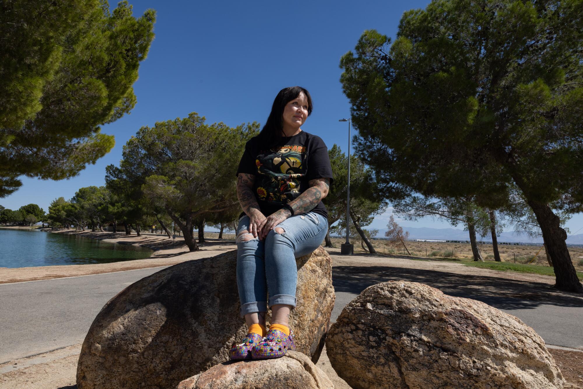 A woman sits on a rock while displaying her purple Lisa Frank Crocs at a park.