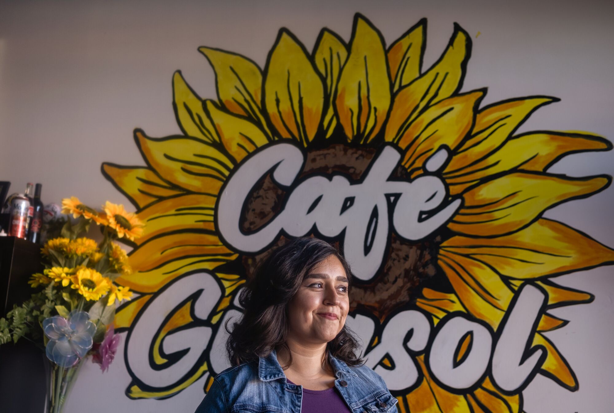 A woman stands in front of a wall on which is painted a sunflower and the words Cafe Girasol.