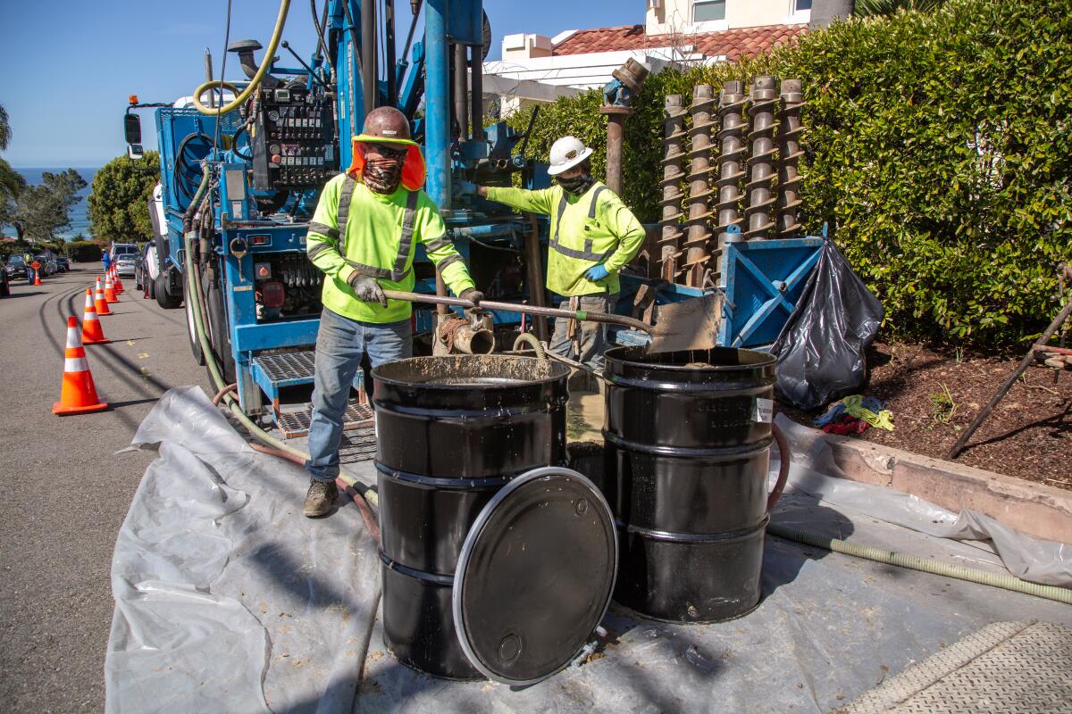 Orange cones are set up alongside workers, drilling equipment and two drum barrels.