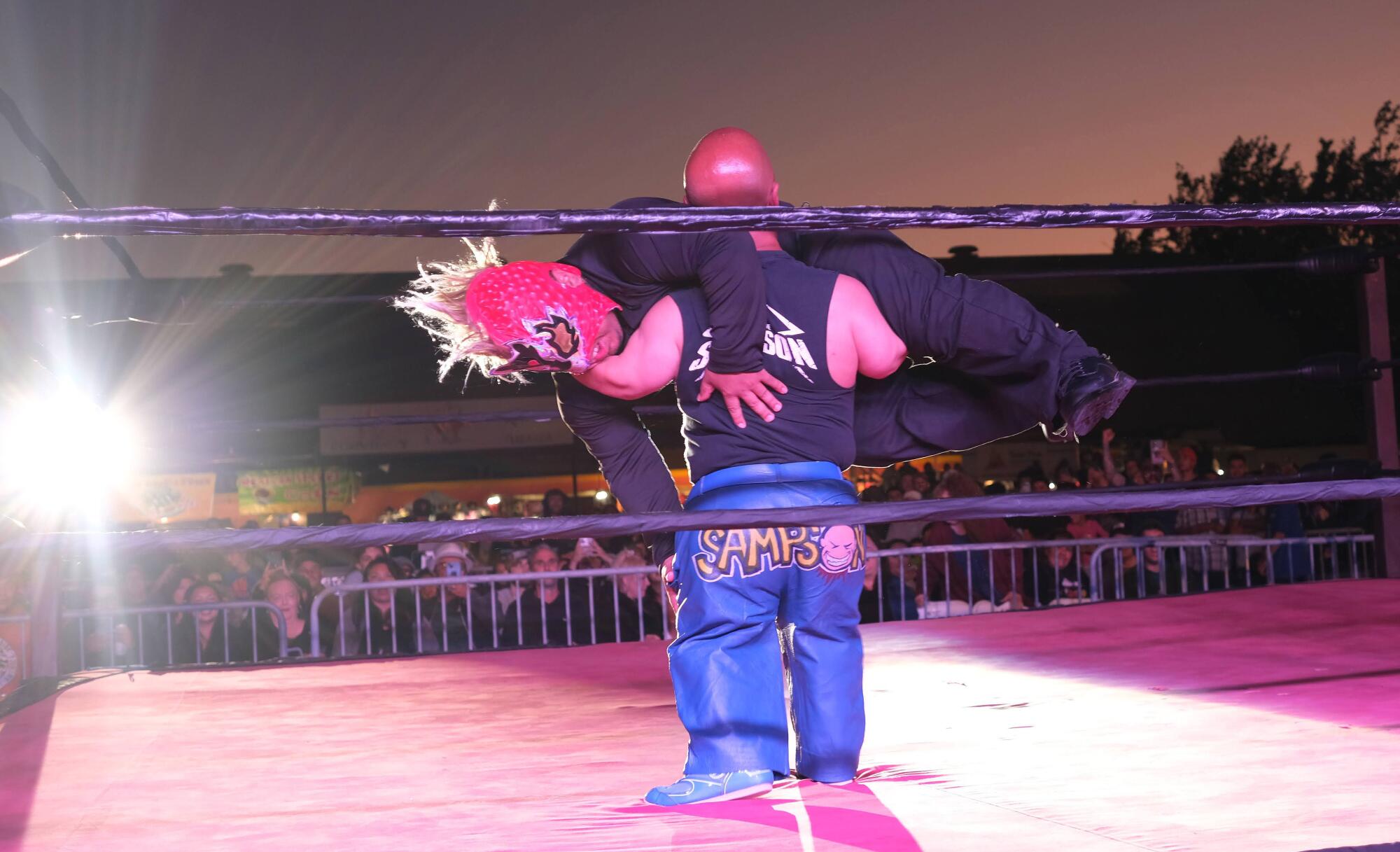 A person holds a horizontal person in a wrestling ring.
