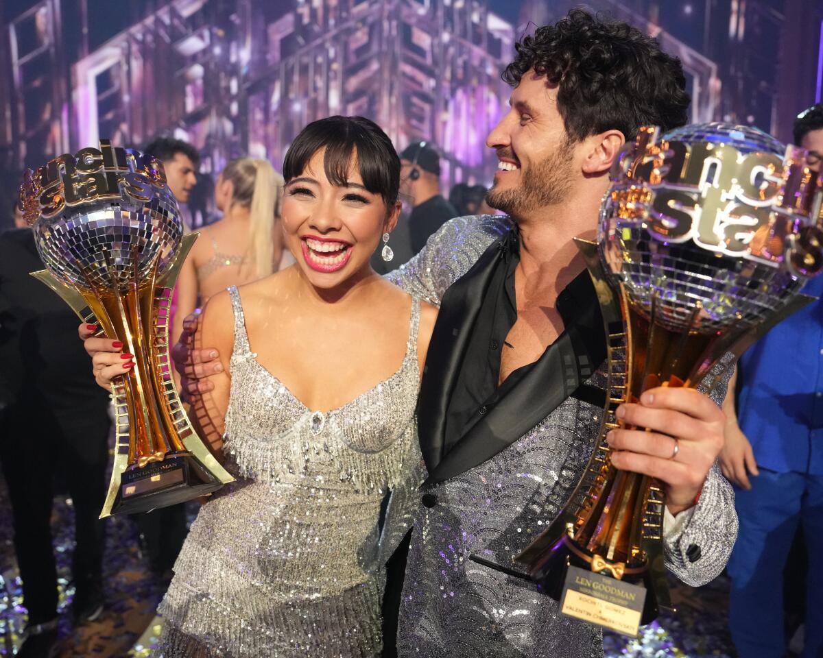Xochitl Gomez in a sparkly silver dress and Val Chmerkovskiy in a silver jacket and black shirt, each holding a trophy.