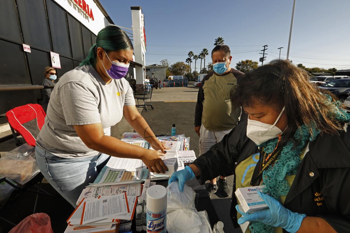 People take masks, pamphlets and boxes of COVID-19 tests from a woman working at a table outside