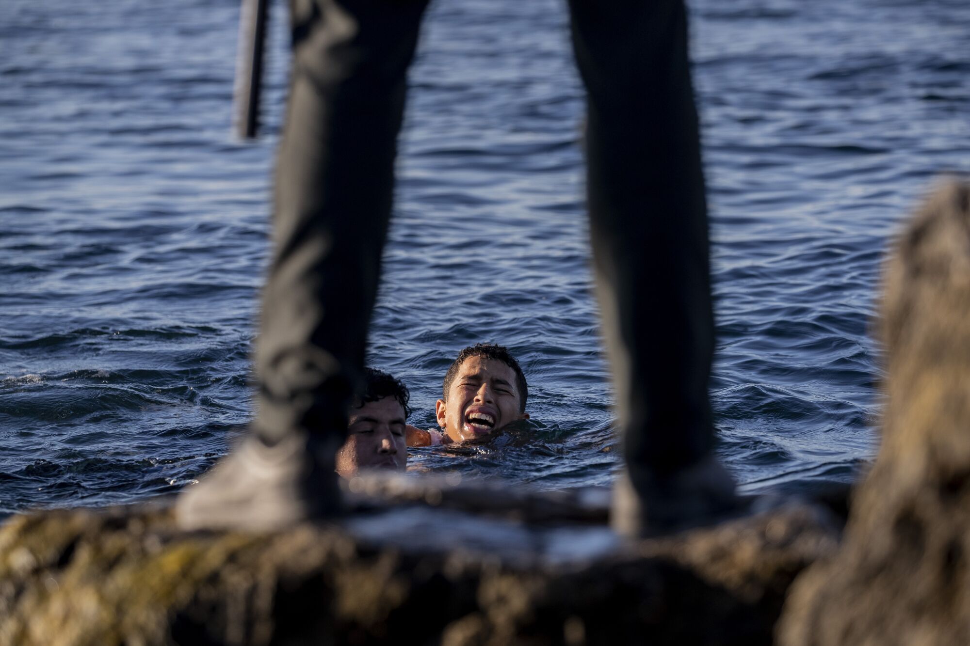 Two young men swimming onto shore are seen between the legs of a police officer standing on a rock