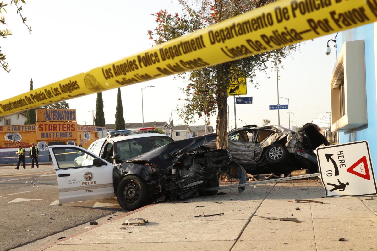 A police car with heavy front-end damage sits next to a tree and downed traffic sign on a sidewalk near a wrecked sedan.
