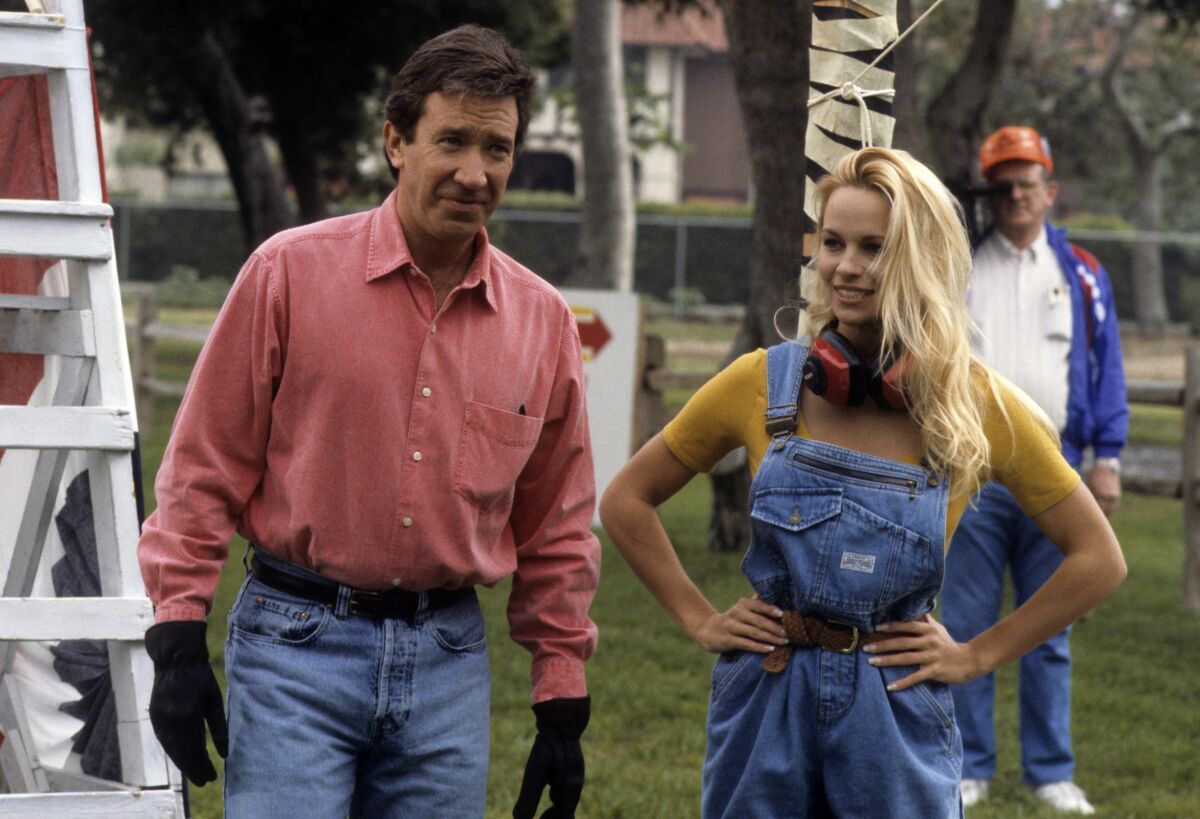 A man in a salmon button-up shirt and jeans stands next to a woman in denim overalls