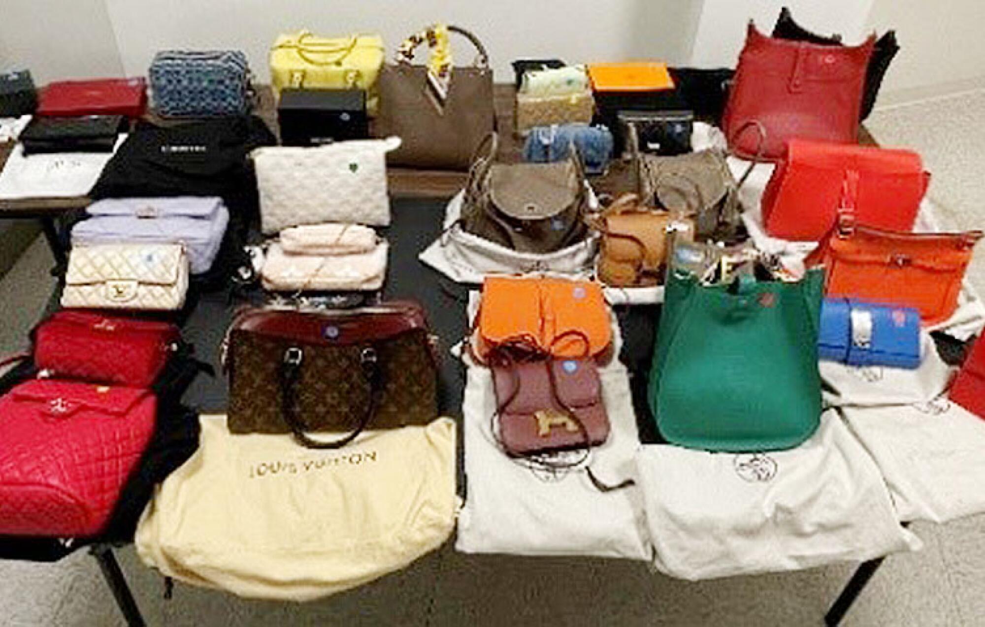 More than two dozen purses of various sizes and colors arranged in rows on a table, some with dust bags from high-end brands