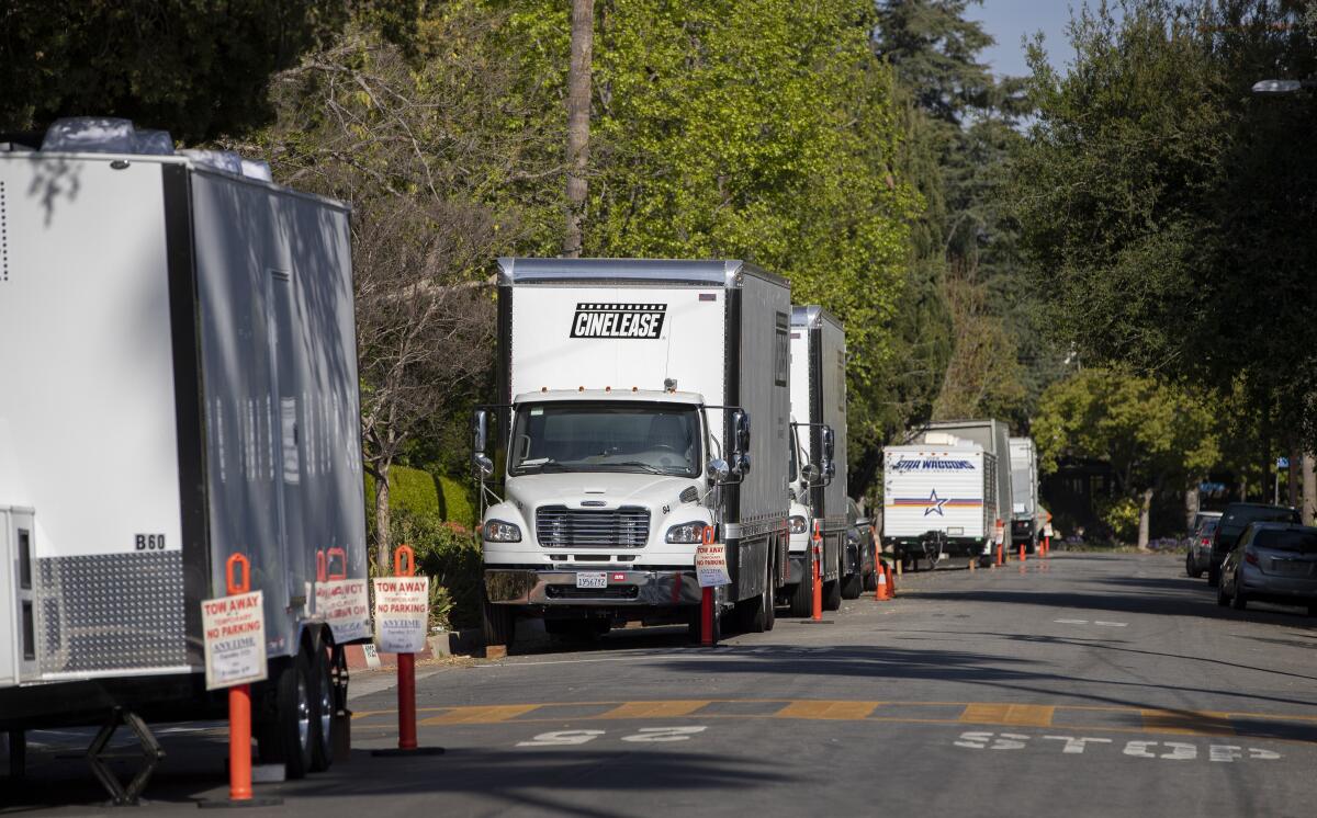 Large white cinema production trucks and trailers are parked along a tree-lined street.