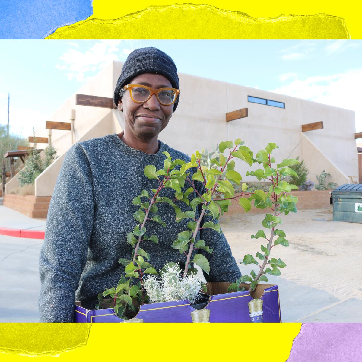 A person in gray sweatshirt, gray beanie and glasses smiles while holding a tray of plants in pots.