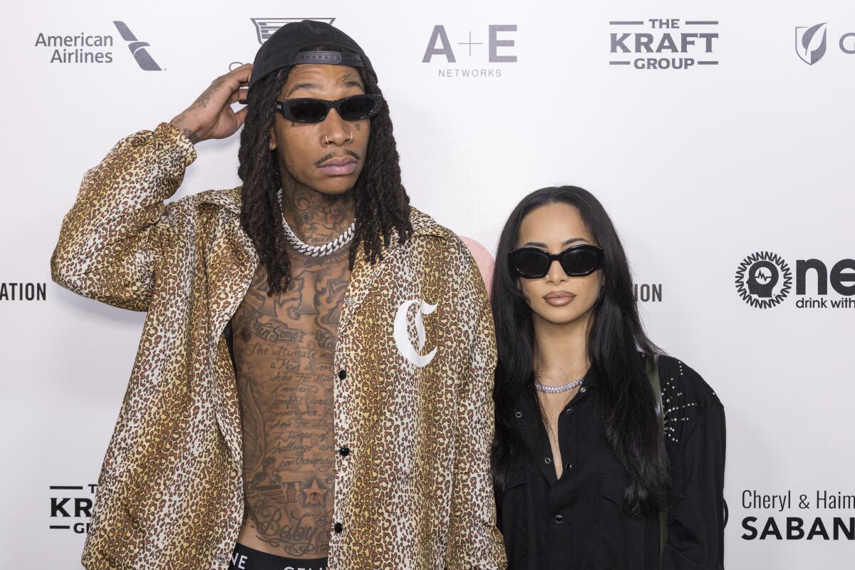 Wiz Khalifa in an unbuttoned cheetah-print shirt and shades standing with Aimee Aguilar in a black top and sunglasses