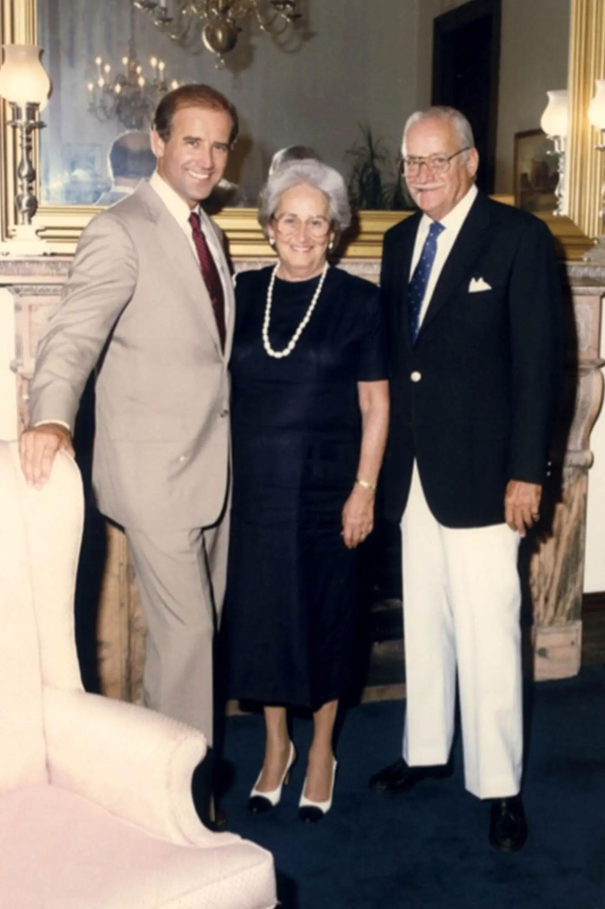 A man in a tan-colored suit and tie, left, stands next to a woman in a dark dress and a man in a dark jacket and white pants 