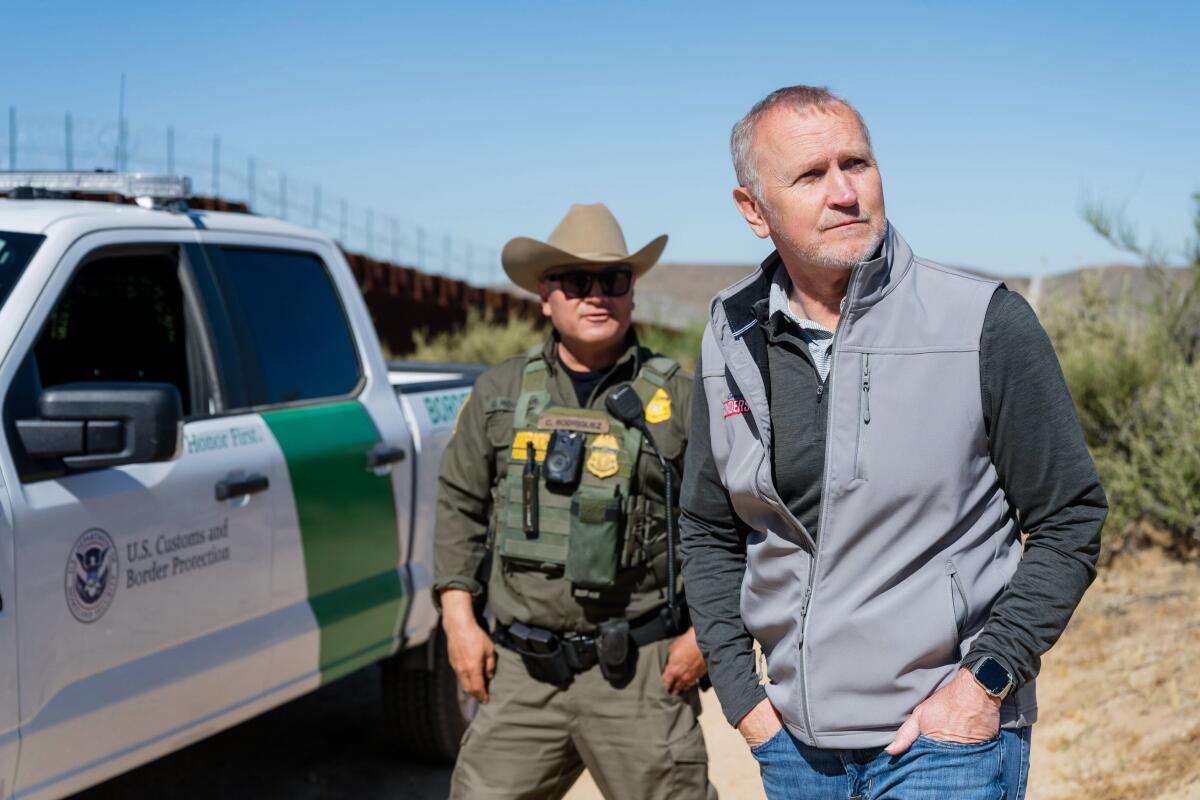 Matt Gunderson y un hombre vestido de verde y un sombrero de vaquero amarillo miran a su alrededor junto a una camioneta blanca del gobierno cerca del muro fronterizo.