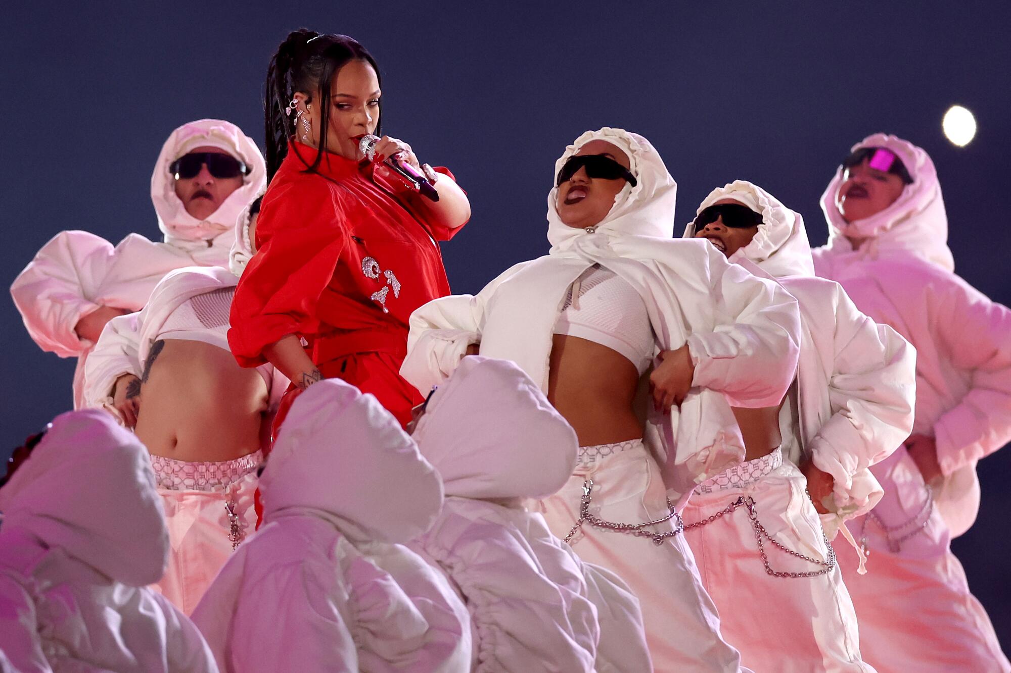 A woman in red sings into a microphone as dancers in white move past her. 