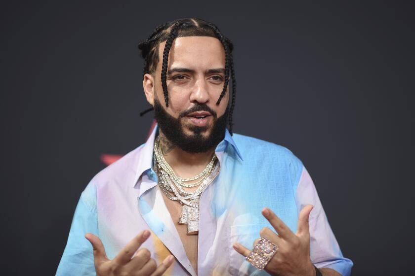 A man with braids, a beard and gold chains over a multicolored shirt makes gestures with both hands