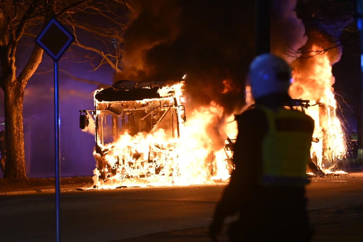 An officer in riot gear looks at a burning city bus.