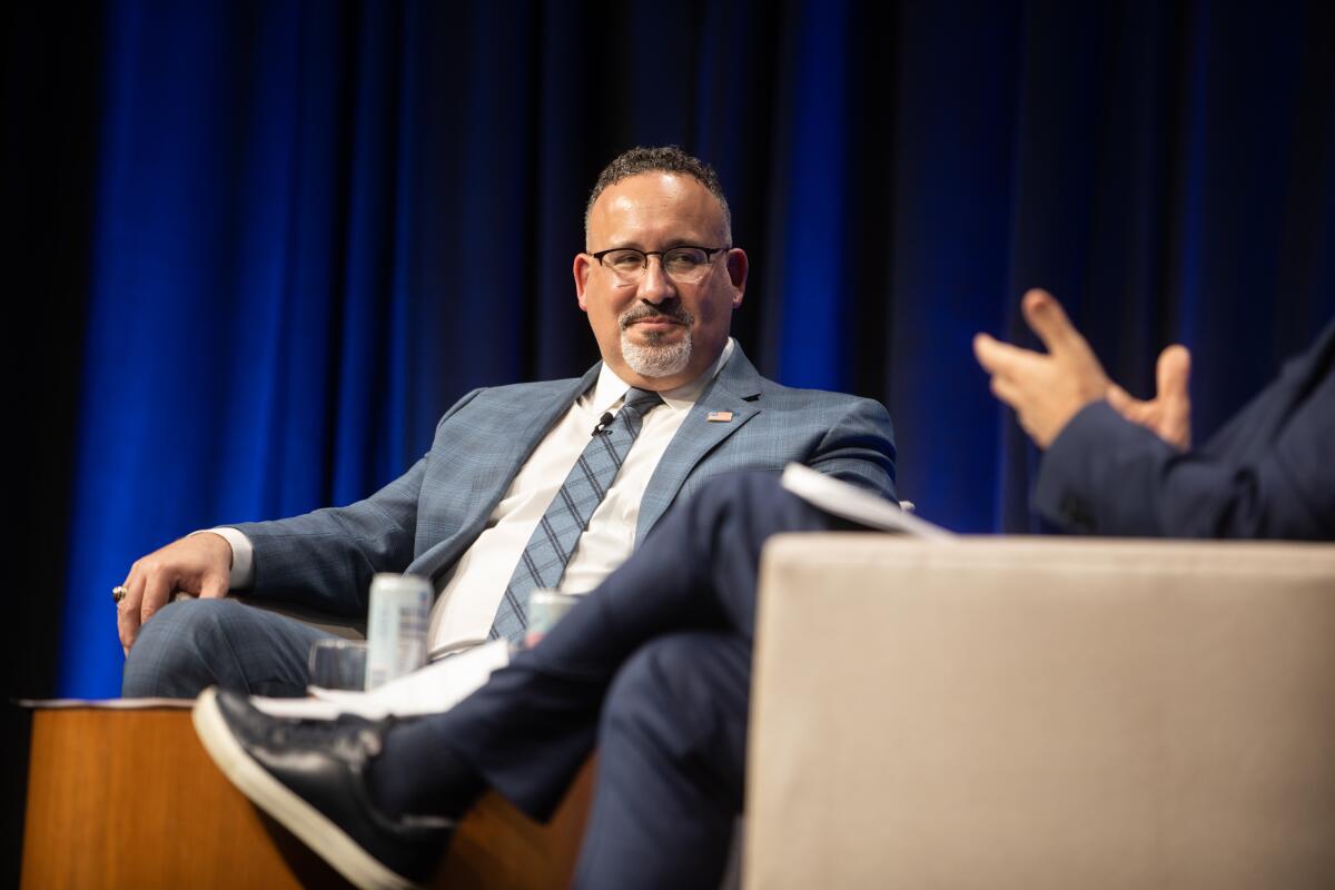 Education Secretary Miguel Cardona  sits on a chair talking. 