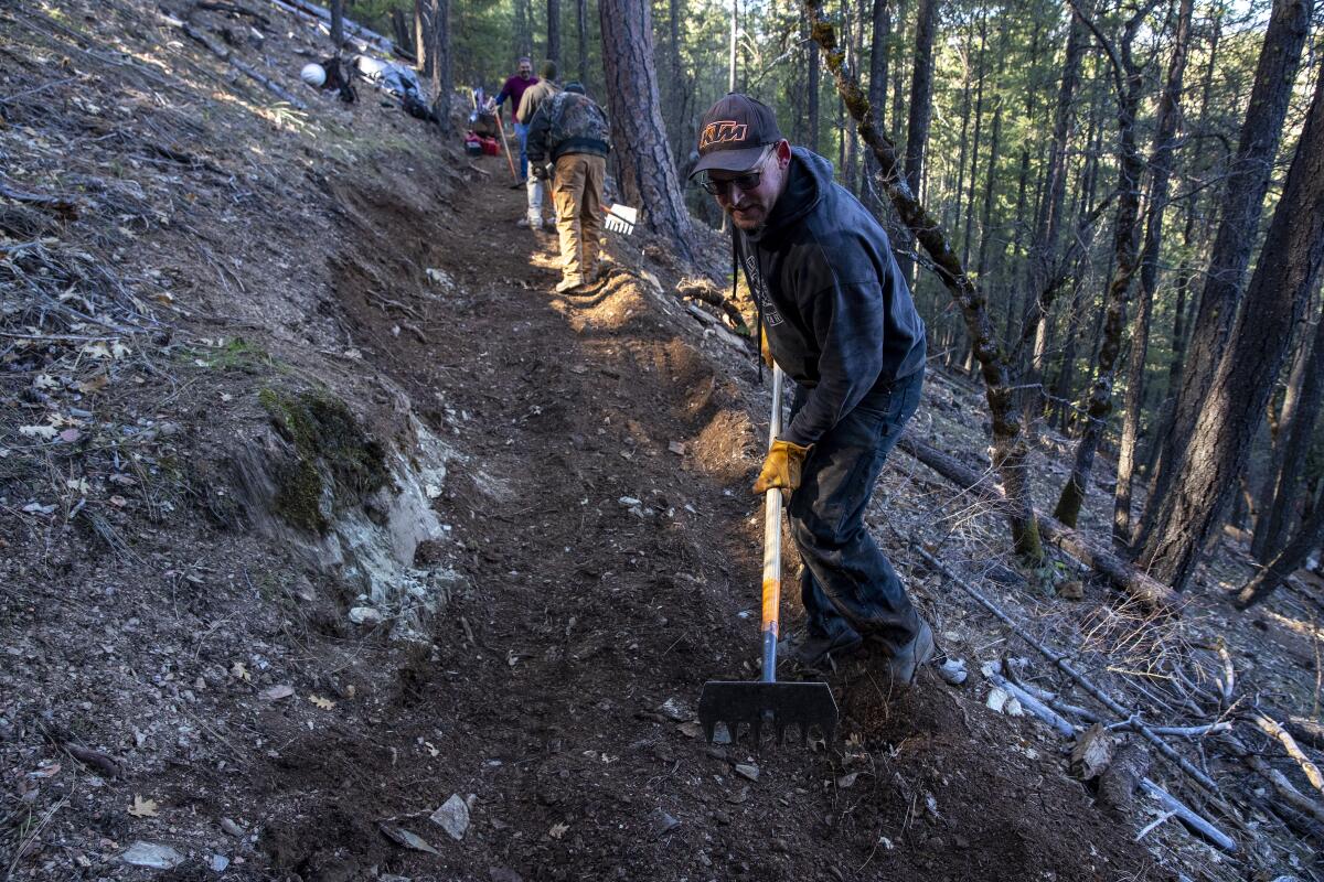 Four people use hoes and rakes on a dirt trail on the side of a forested hill.