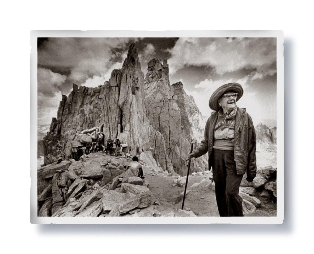 A woman with a hat and walking stick smiles while standing on a mountain trail.