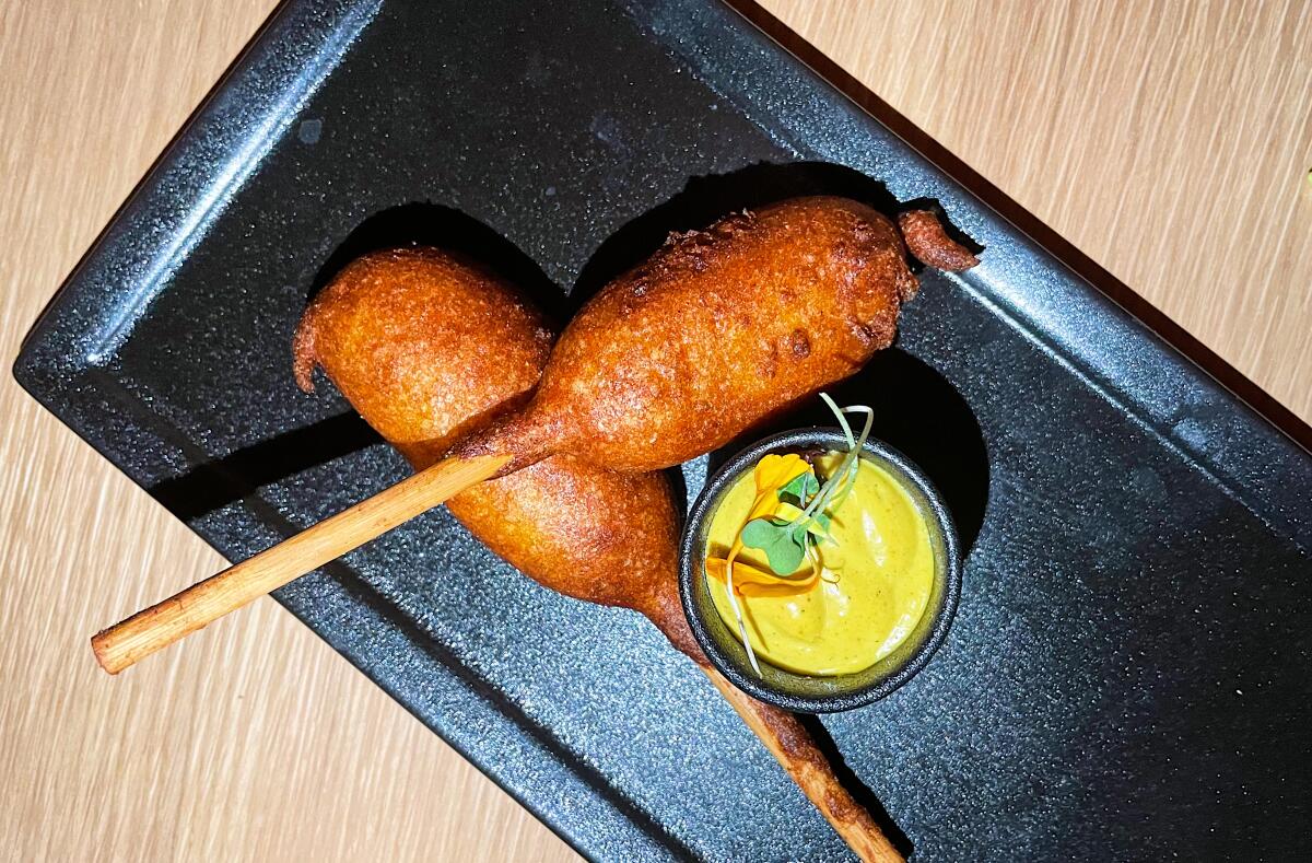 An overhead photo of small corn dogs on sticks on a black plate with a small container of yellow sauce