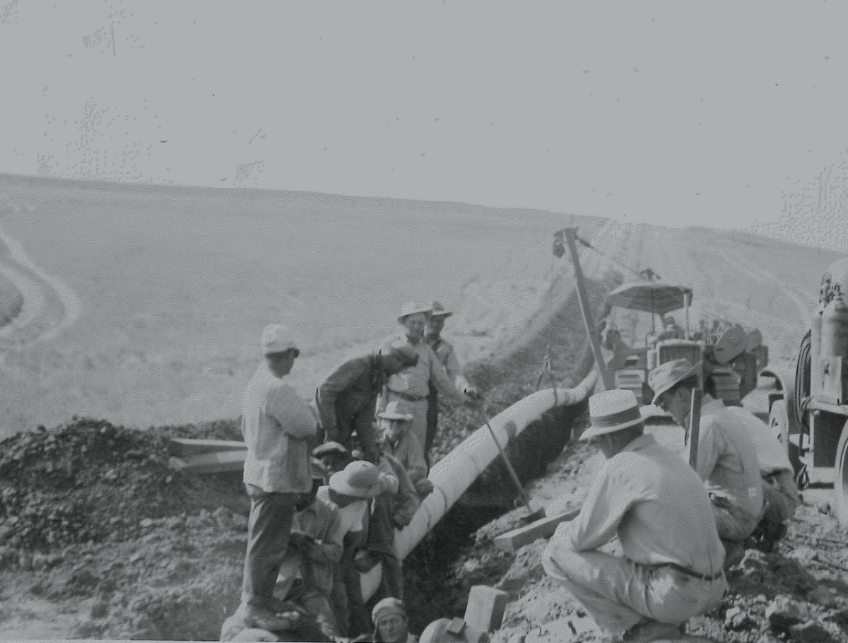 Black and white photo shows men working on a pipeline in a rugged landscape.