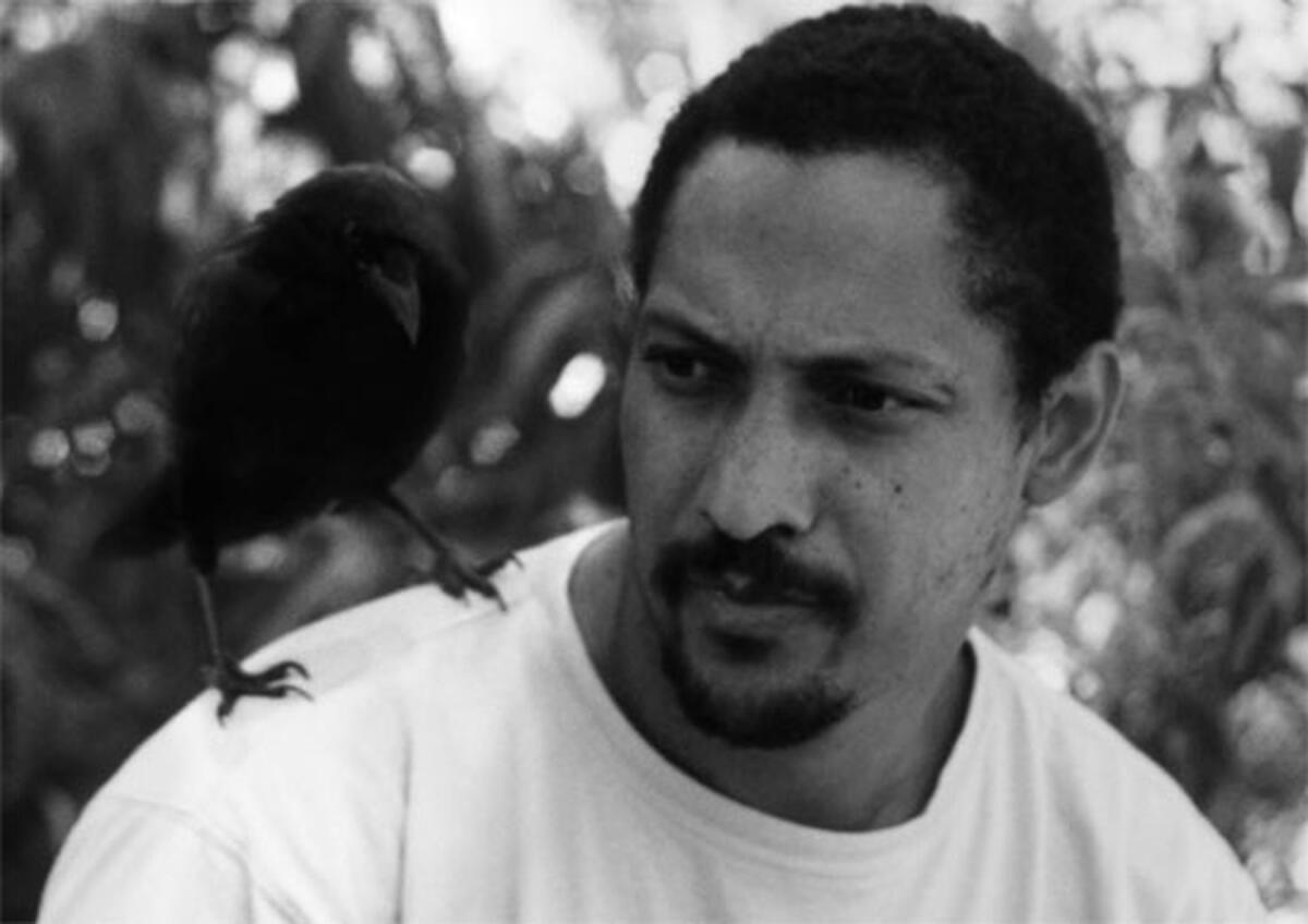 Closeup of a man with a goatee and wearing a white T-shirt; a bird is perched on his shoulder.