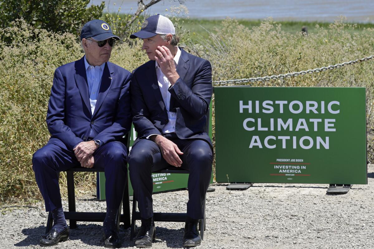 Gov. Gavin Newsom talks with President Biden on a bench surrounded by greenery 